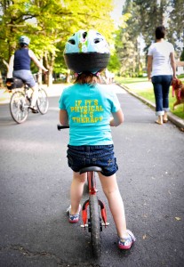 Little girl on bike seen from te back, wearing helmet, others biking/walking in street.