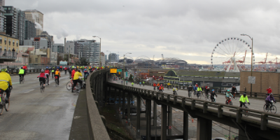 People on bikes ride across the now-defunct SR 99 tunnel with the Seattle Ferris Wheel in the background