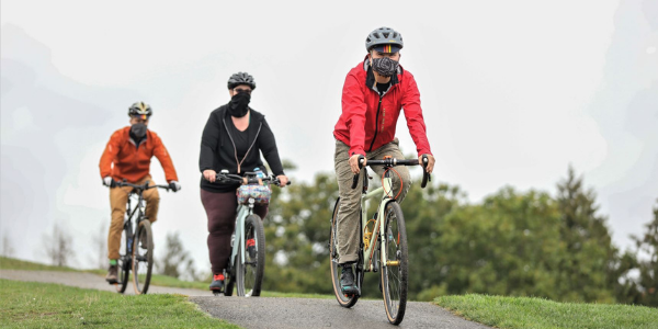 Three people ride bikes (a Latino man in a red jacket, a large-bodied woman in a black jacket, and a white man in an orange jacket).