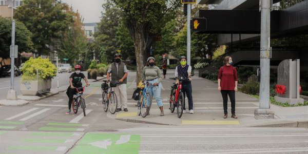 A Black child, Black man, Black woman, and Asian man stand next to their bikes beside a white woman walking. All are waiting to cross an intersection while wearing masks.